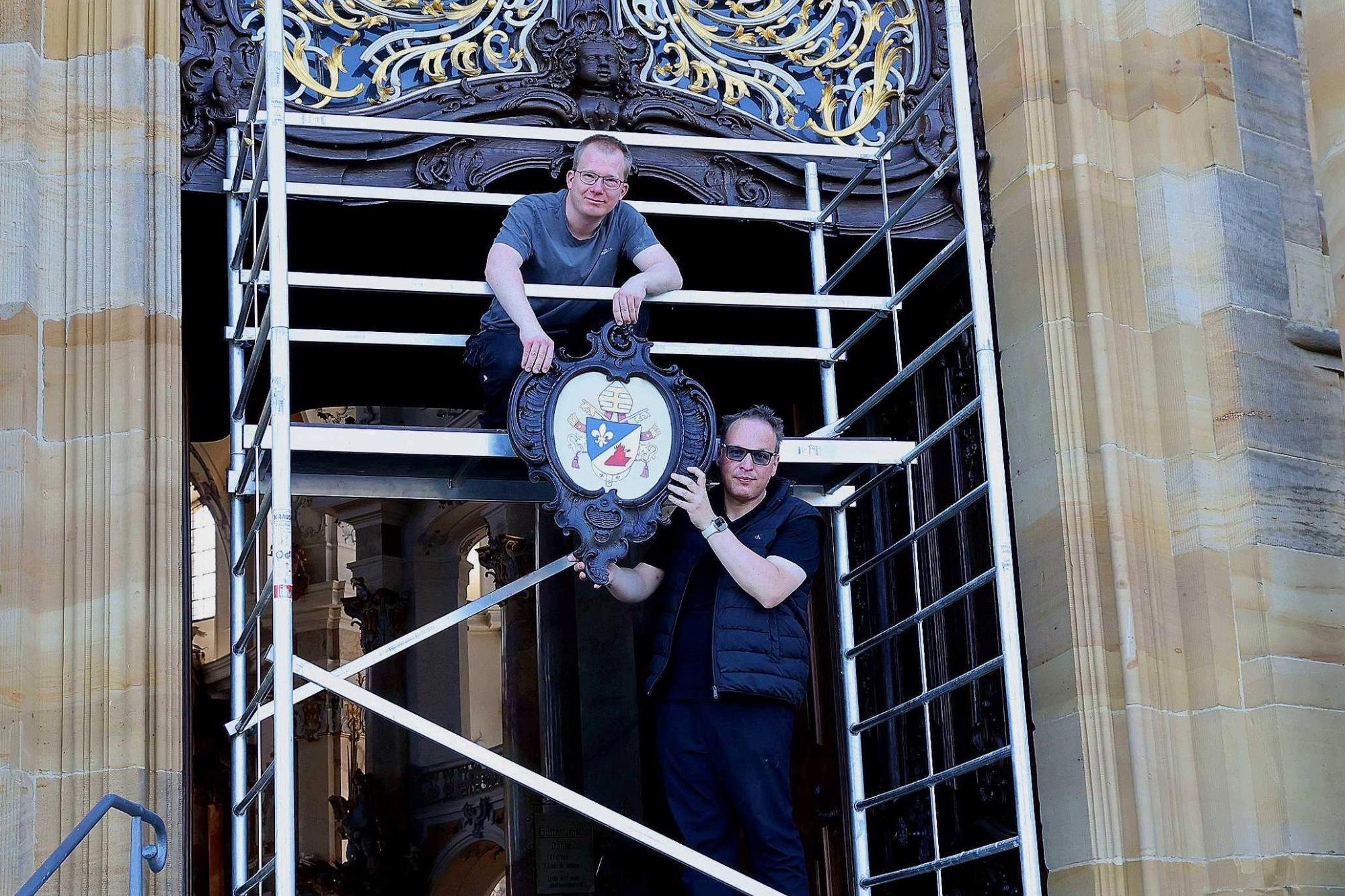 Basilikamesner Tobias Hartmann und Kirchenschweizer Daniel Reitz bringen gerade das neue Papstwappen am Hauptportal der Wallfahrtskirche an.