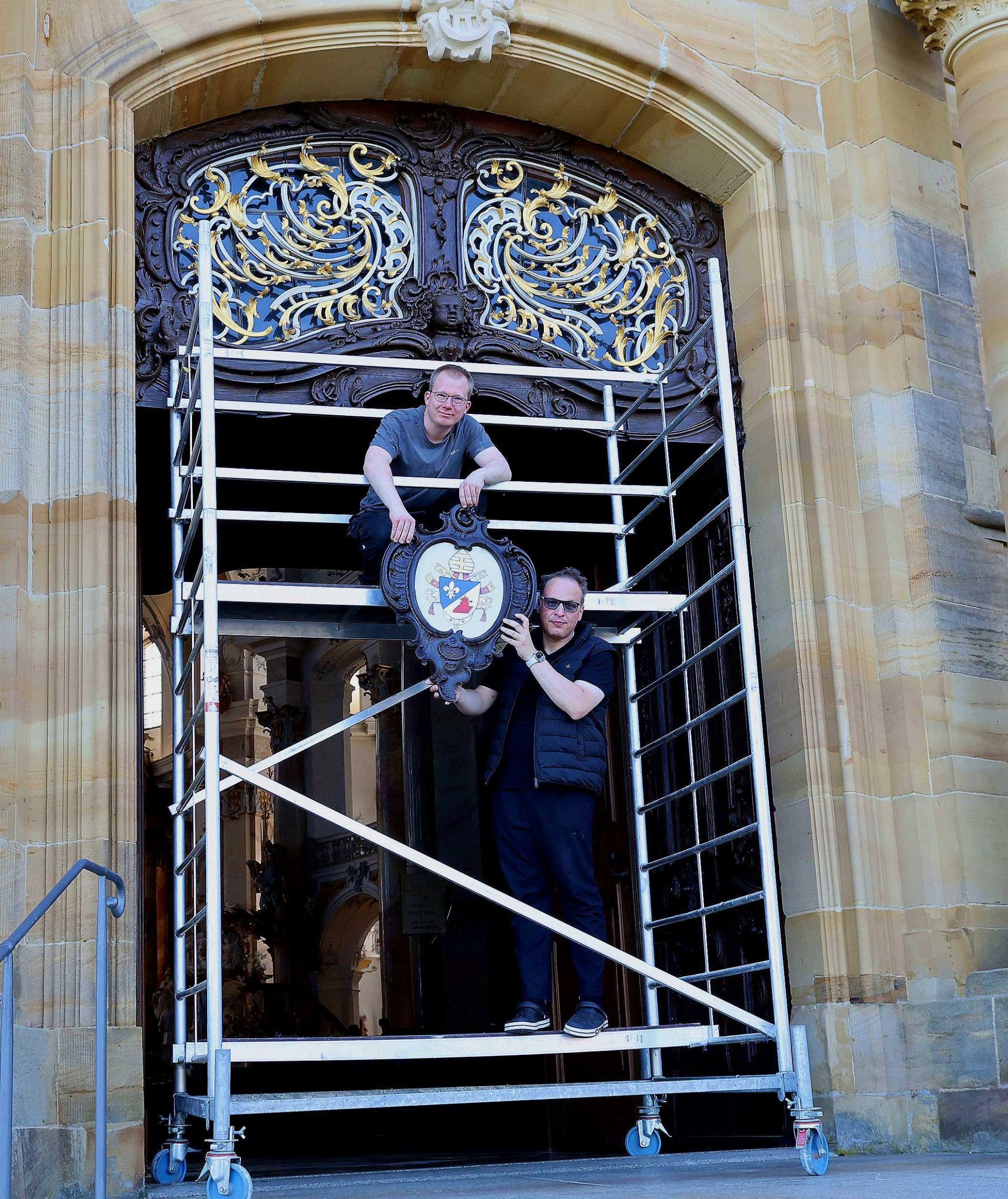 Basilikamesner Tobias Hartmann und Kirchenschweizer Daniel Reitz bringen gerade das neue Papstwappen am Hauptportal der Wallfahrtskirche an.